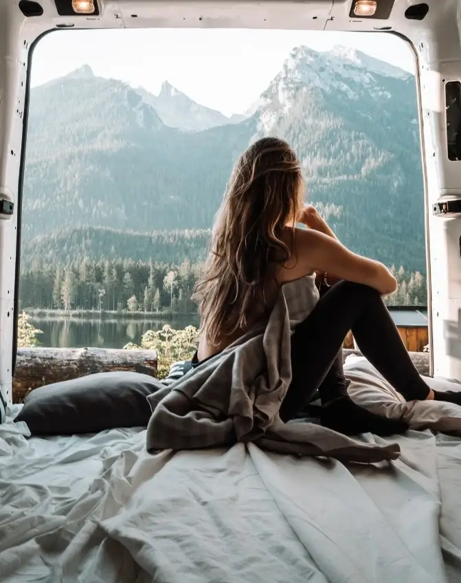A woman sitting in the converted cargo space of her van on a mattress and staring out the back at a mountain landscape.
