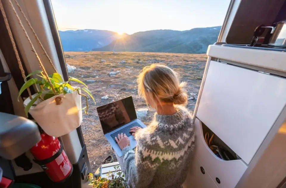 A girl sits in a well-maintained panel van, working on her computer while watching the sun set over the mountains,.