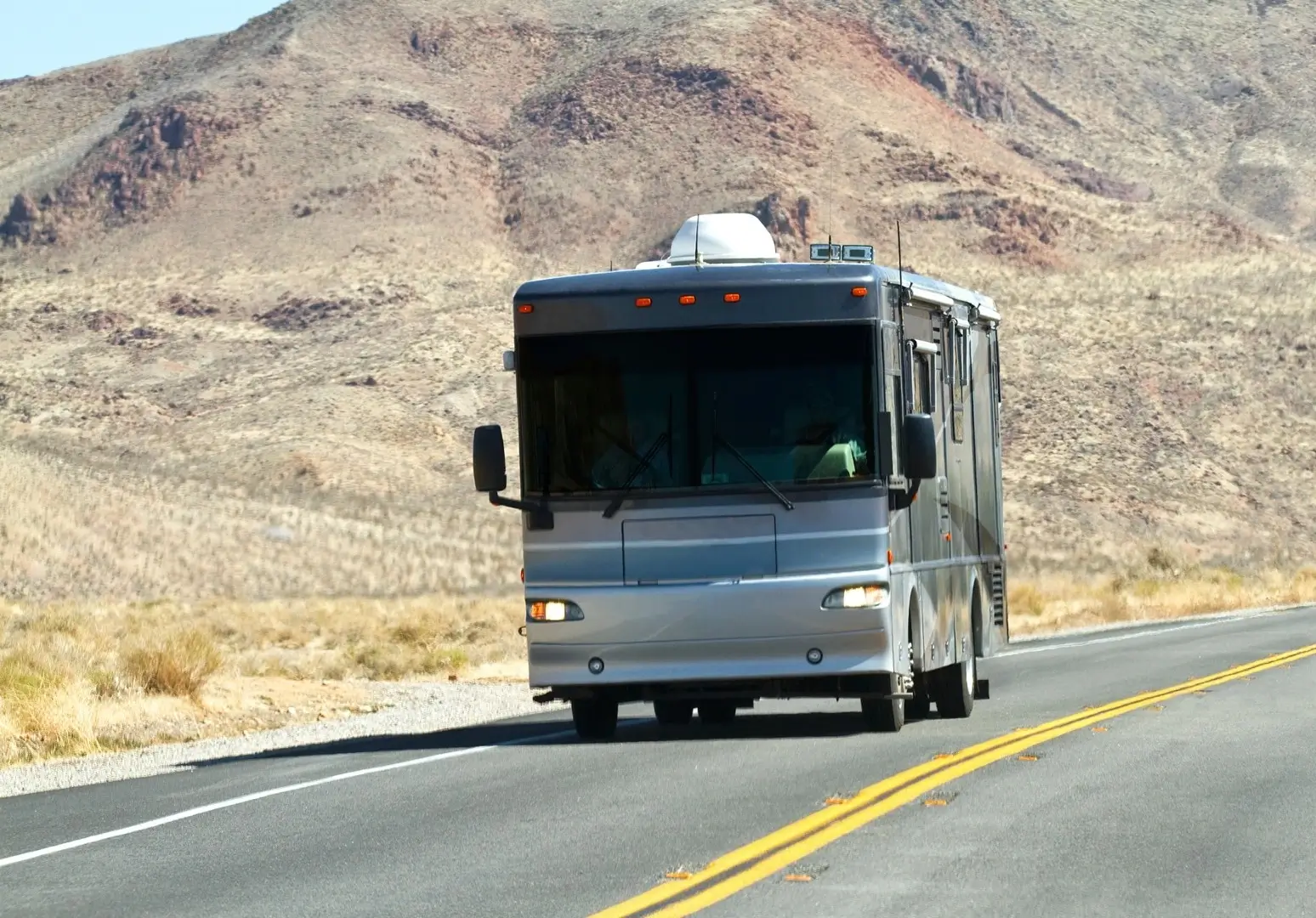 A class A motorhome travels down a desert highway with mountains in the background.