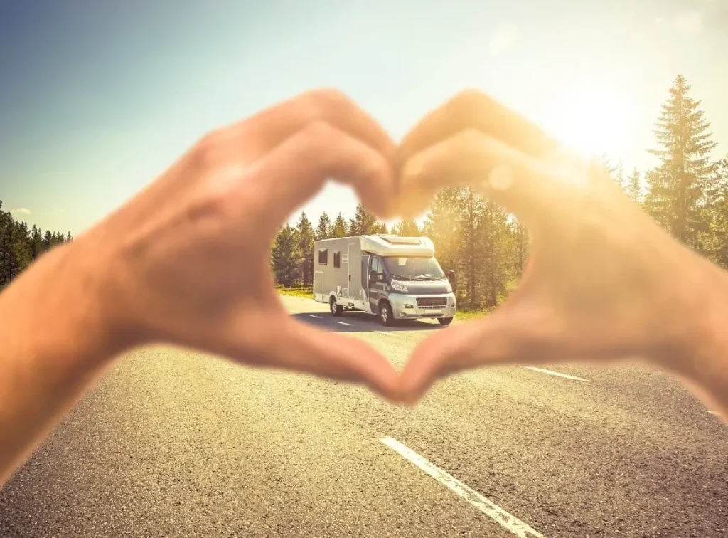 A woman forms a heart shape with her hands around her Class B motorhome, parked on a tree-lined street.