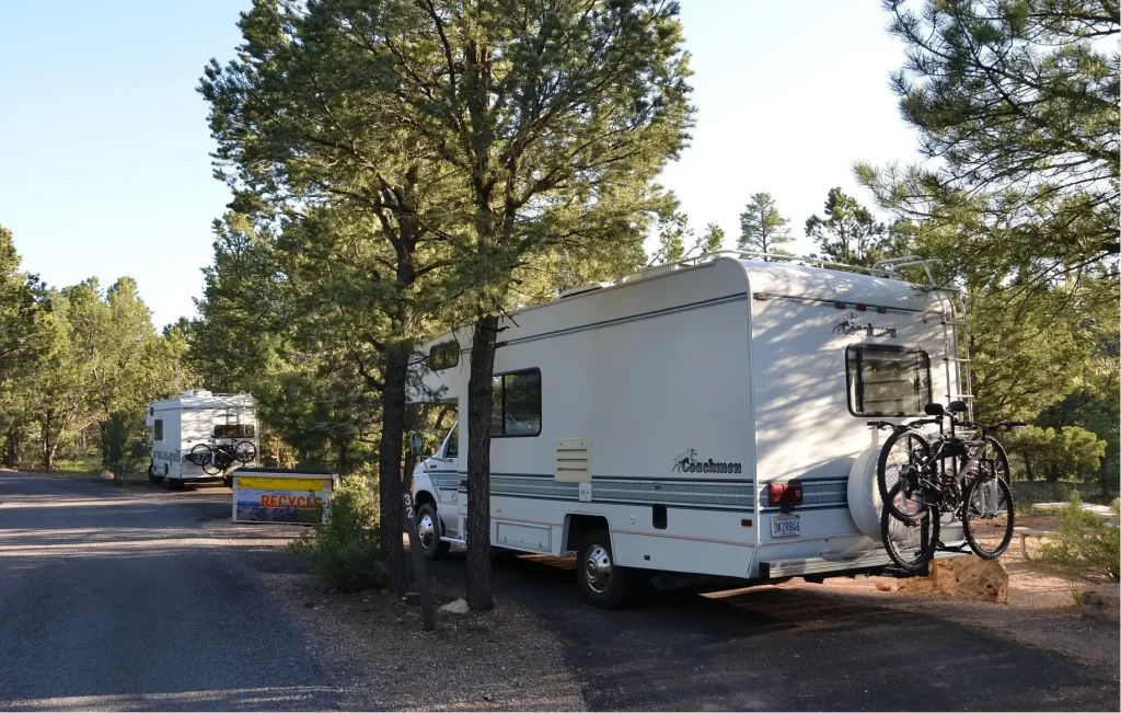 An affordable Coachmen motorhome is parked by the roadside near a campground, its bumper loaded with bikes.