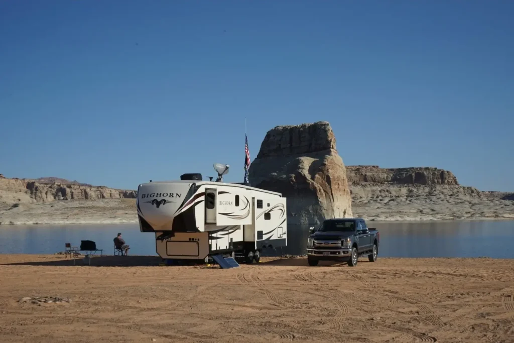 A versatile Heartland fifth-wheel travel trailer is parked beside a pickup truck on a desert lakeshore, with a towering rock formation in the background.