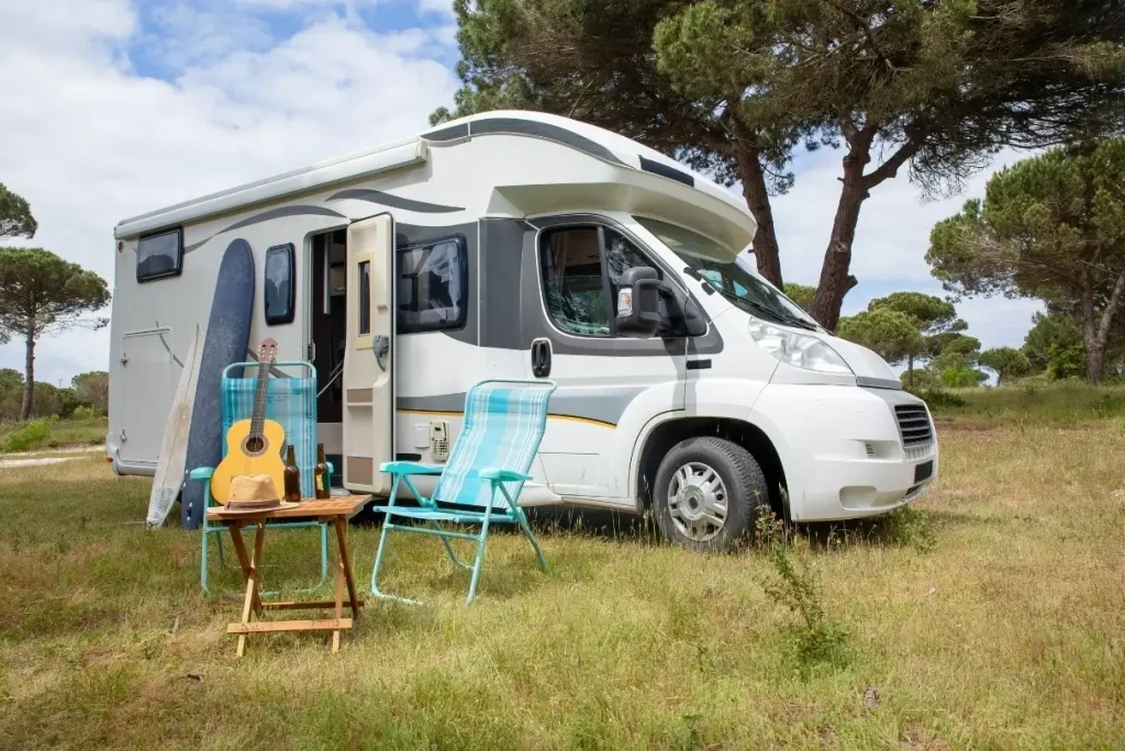 A fully kitted out Class C motorhome camped out beneath trees with a collection of its must-have RV essentials spread out in front.