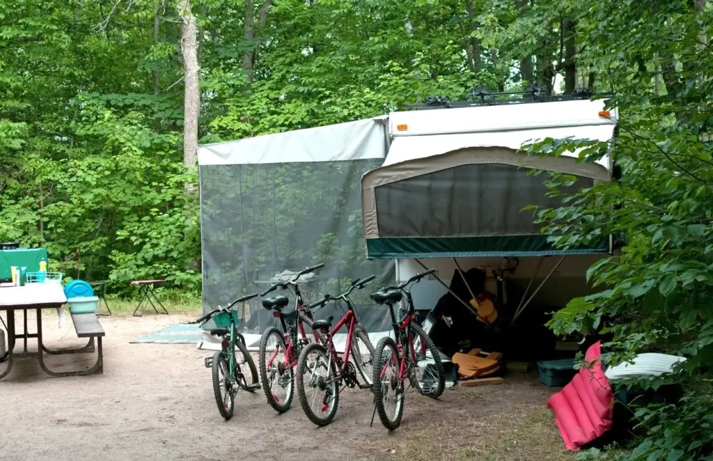 This pop-up camper sits parked in a campground next to a picnic table, with several bikes and a pool float.