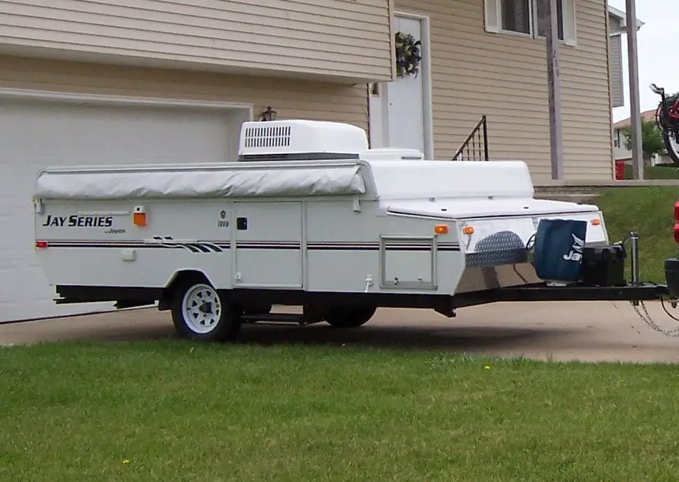 A Jayco pop-up camper sits folded down and hooked up to the tow vehicle in front of a family home.
