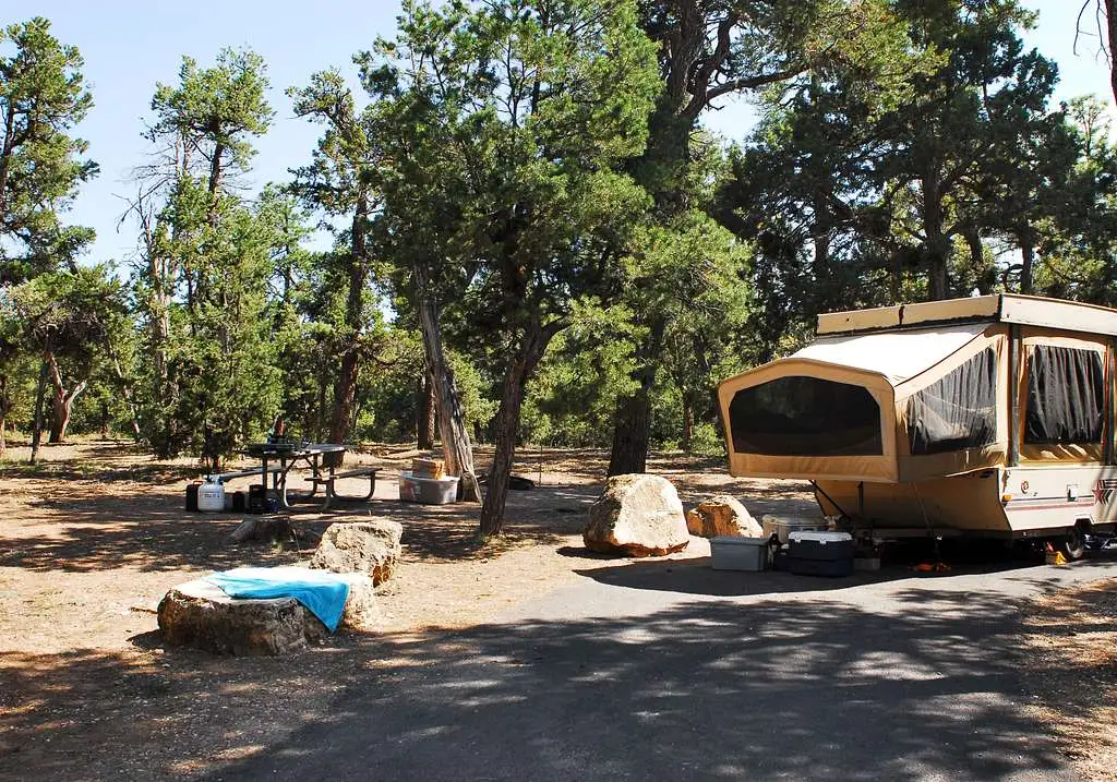 A tan-colored pop-up camper is parked at a campsite under tree cover, with large boulders surrounding it.