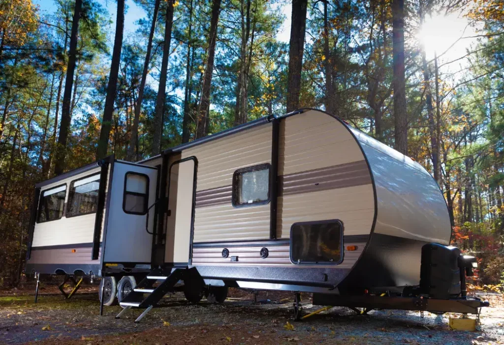 A tan travel trailer with a light brown stripe and an extended slide sits in a forest with tall trees and sunlight shining through.
