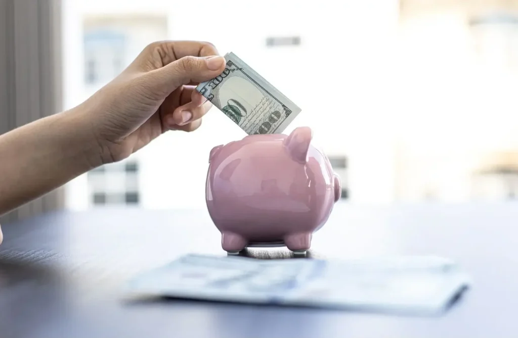A person inserting a $100 bill into a pink piggy bank on a table.