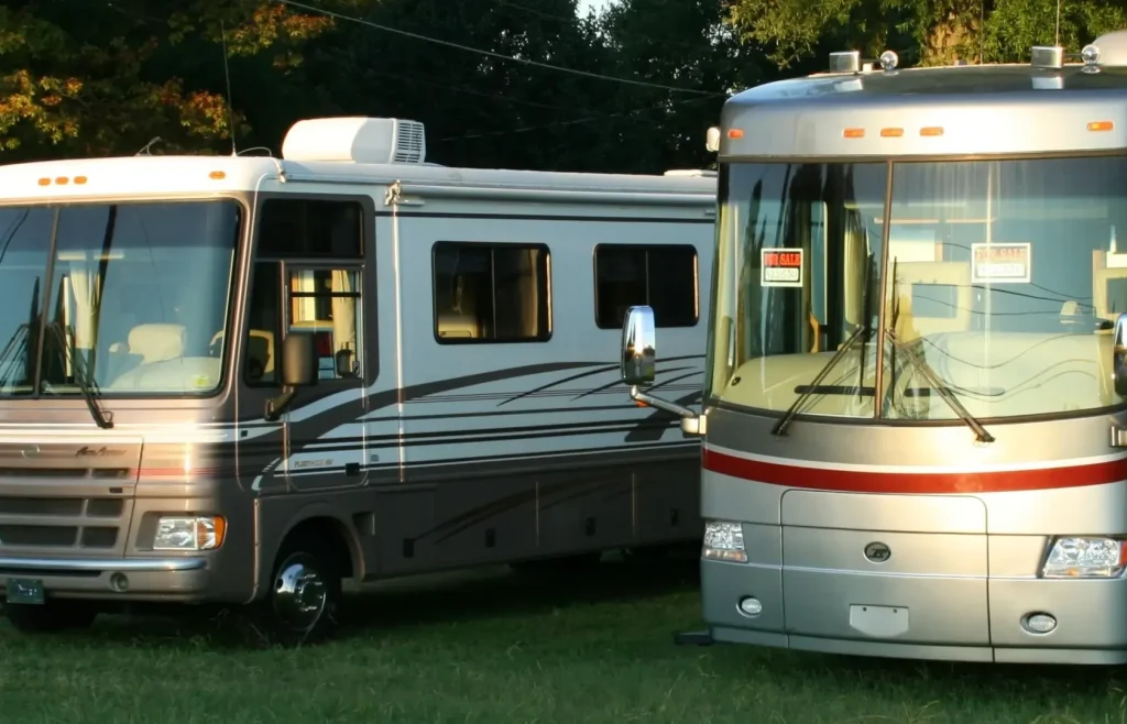 Two older model RVs are parked in a grassy lot with 'For Sale by Owner' signs displayed in the windows.